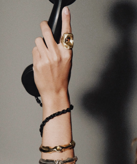 Mão feminina segurando gancho de telefone antigo preto, usando anel dourado grande de formato orgânico com pedra central e braceletes metálicos no pulso. Mão feminina segurando gancho de telefone antigo preto, usando anel dourado grande de formato orgânico com pedra central e braceletes metálicos no pulso.