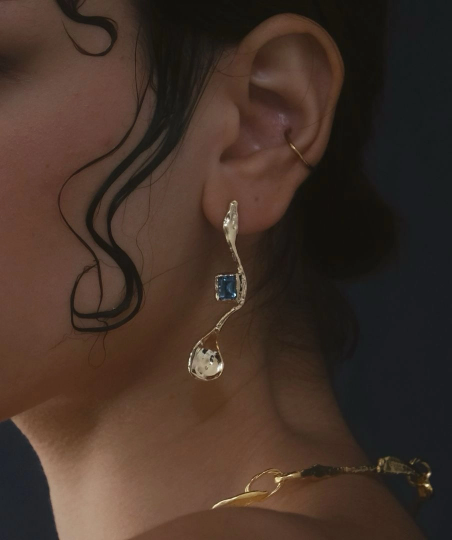 Close de brinco longo dourado de formato fluido e orgânico com pedra retangular azul incrustada e piercing de argola fina na orelha. Close de brinco longo dourado de formato fluido e orgânico com pedra retangular azul incrustada e piercing de argola fina na orelha.