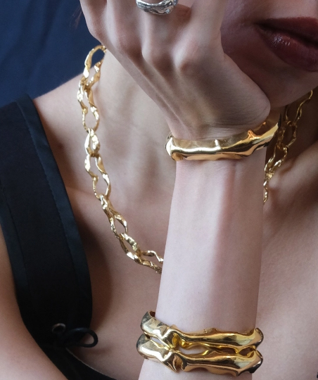 Close de modelo usando conjunto de joias douradas de design orgânico: colar de elos texturizados, braceletes espessos nos pulsos e anel de prata na mão apoiada no queixo.