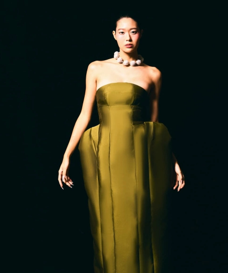Modelo usando vestido longo tomara que caia tom verde oliva acetinado, com silhueta escultural volumosa no quadril e colar de pérolas esféricas em fundo preto.