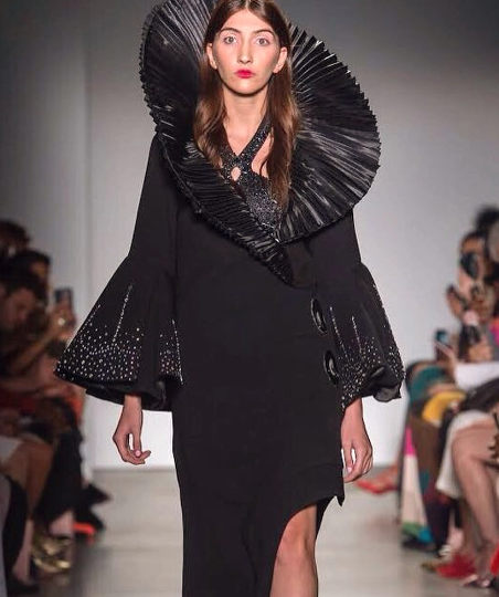 Modelo desfilando com vestido preto conceitual de gola plissada gigante ao redor do pescoço, mangas sino com bordados em pedrarias e fenda na saia.