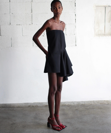 Modelo usando vestido curto tomara que caia preto em alfaiataria assimétrica com drapeado lateral e salto vermelho.